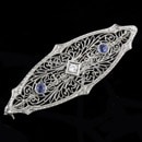 A 14kt white gold diamond and natural sapphire pin from the Edwardian Era, 1901 to 1915. The pin is set centered with one European cut diamond weighing .05ct and flanked by two 2.3mm old cut blue sapphires.  This pin weighs 2.8 grams and is 1 11/16th" x 15mm. This old gal is in pristine condition and a great example of the Edwardian Era.